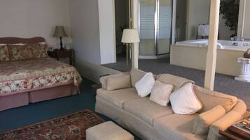 A bedroom with a bed, a sofa, a small table, a lamp, and a bathroom visible through an open door.