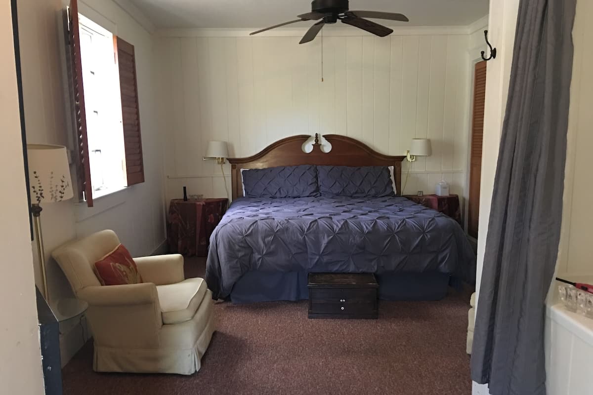Un dormitorio con cama, una silla, una mesita de noche, un ventilador de techo y una ventana.