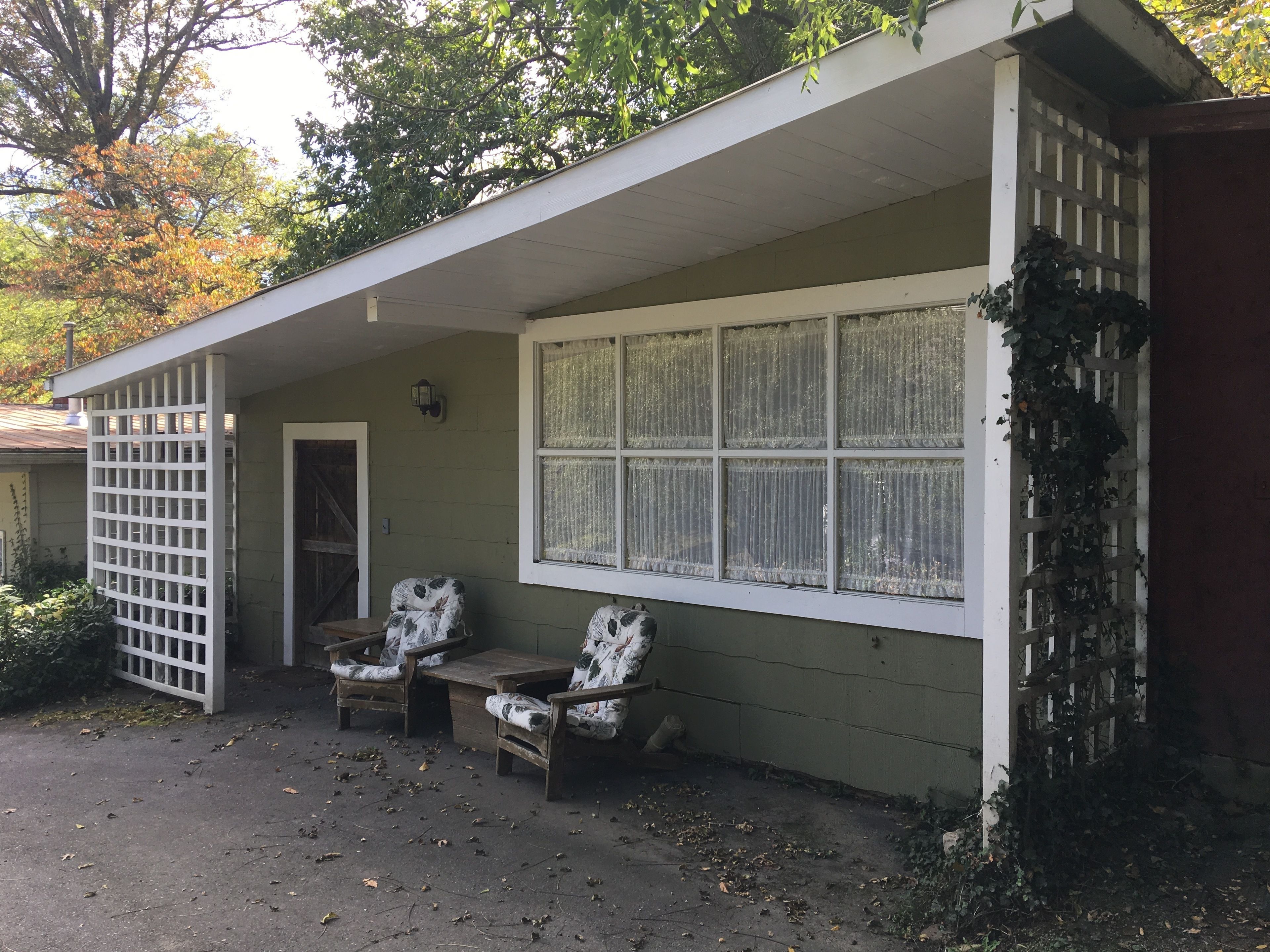 A small building with a porch, two wooden chairs, a table, and a window with lace curtains.