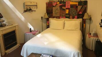 A hotel room with a large bed, a small table with a magazine, a lamp, a bookshelf, and a wall-mounted heater.