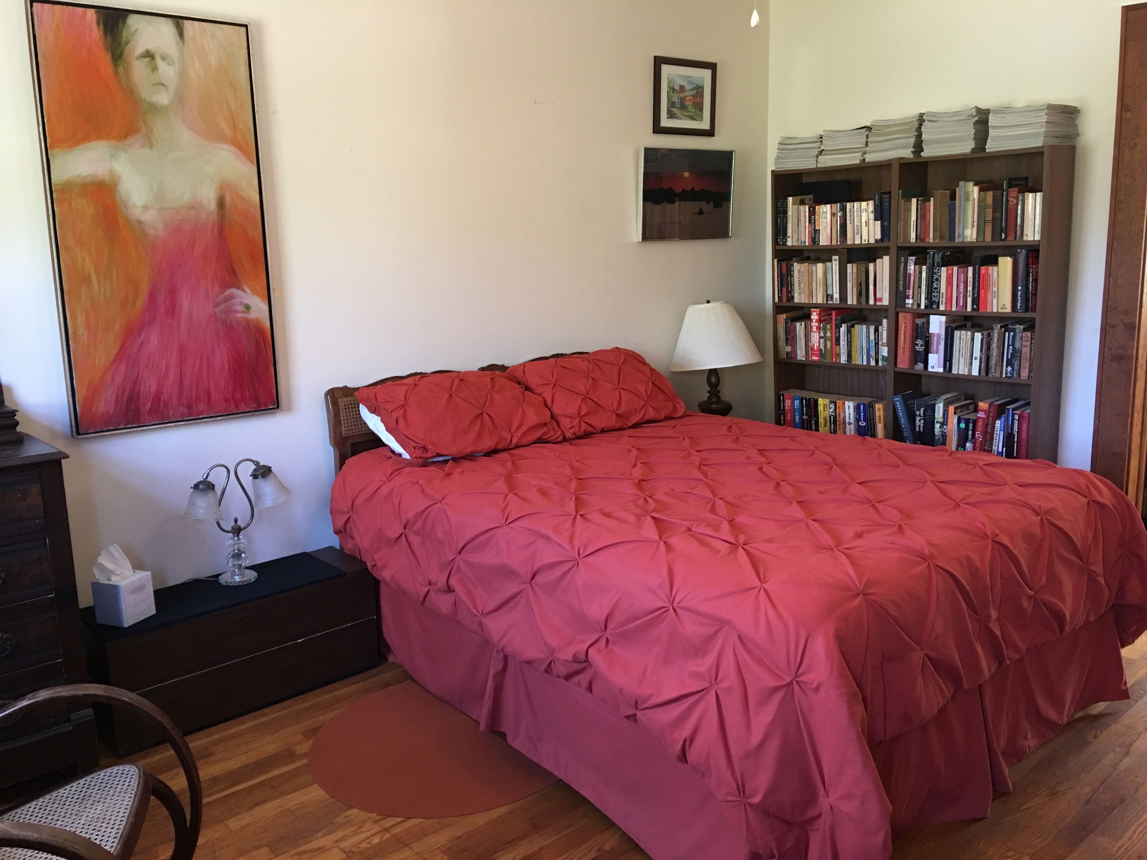 Un dormitorio con una colcha acolchada roja, una mesita de noche con lámpara, una estantería llena de libros y un cuadro en la pared.