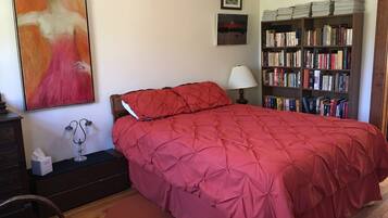 Un dormitorio con una colcha acolchada roja, una mesita de noche con lámpara, una estantería llena de libros y un cuadro en la pared.