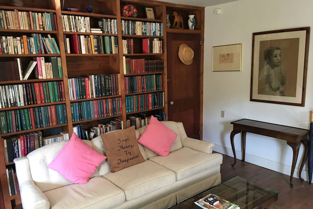 Una sala de estar acogedora con un sofá color beige, dos almohadas rosas y una mesa de centro de vidrio con una revista. Un estante de madera lleno de libros se ve al fondo.