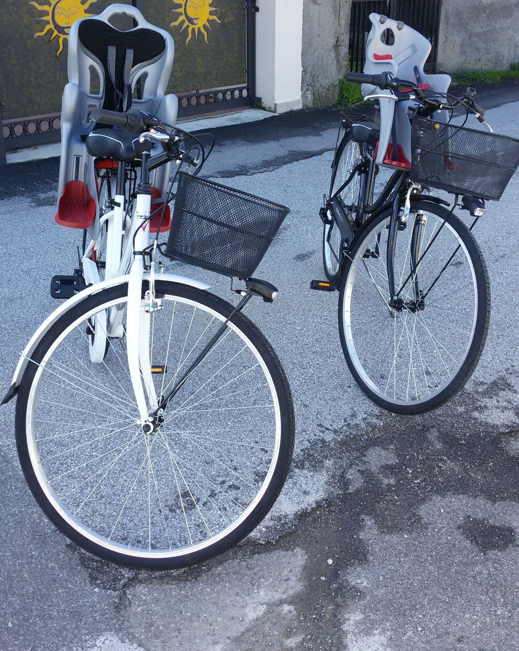 Två cyklar står parkerade på en asfalterad yta, med en byggnad i bakgrunden.
