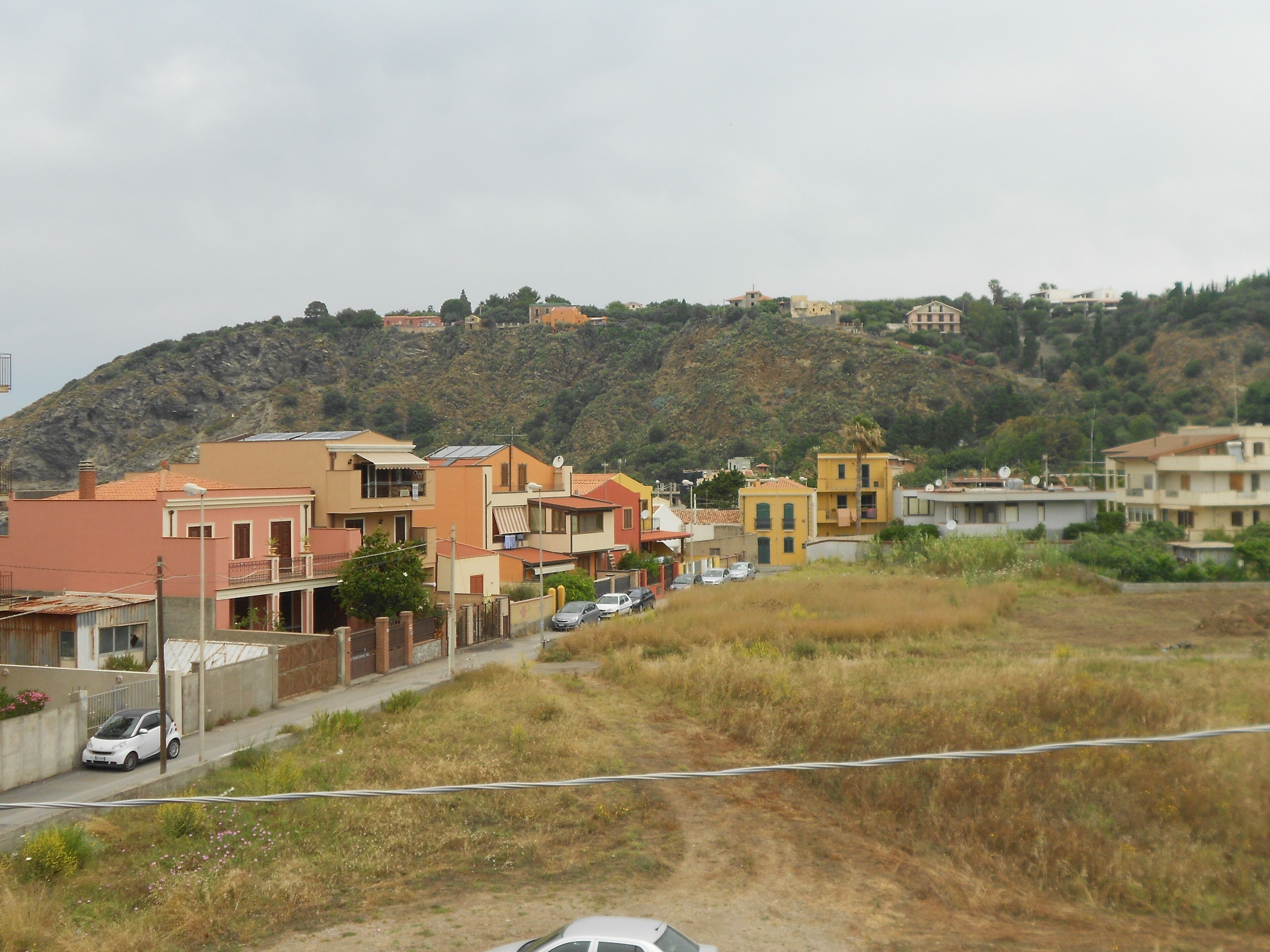 Una strada residenziale con case, un sentiero sterrato e uno sfondo collinare.