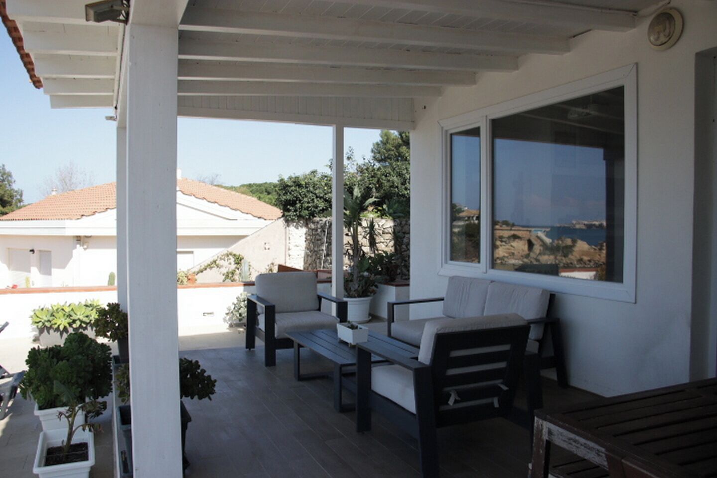 A covered outdoor seating area with a view of the sea, a white wall, and a window.