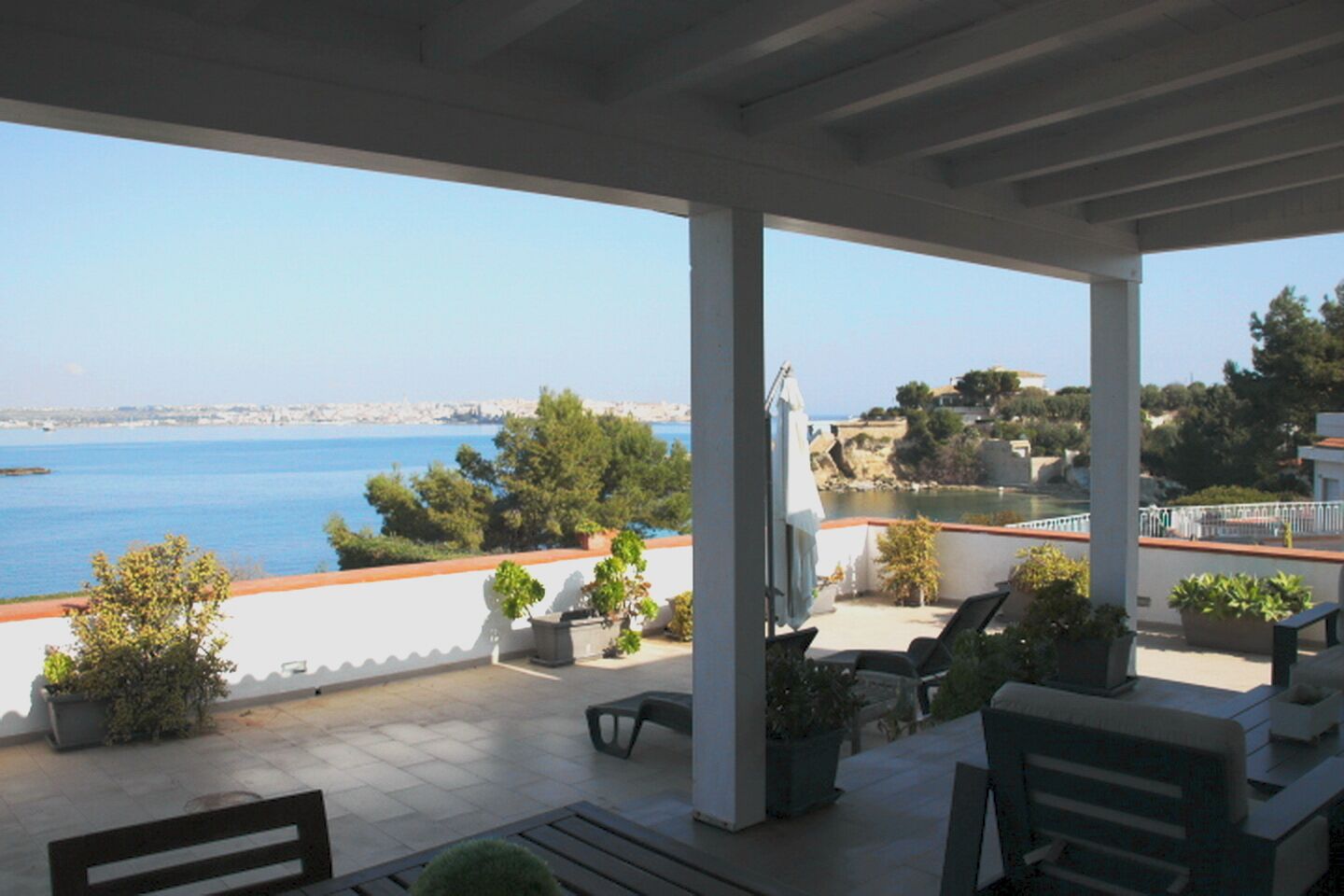 A covered outdoor area with a view of the sea, seating, and potted plants.