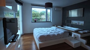 A modern bedroom with a large bed, a TV, a window with a view of greenery, a built-in sink, and a mirror. - design villa on the sea with a splendid view of the Gulf of Syracuse (Siracusa)