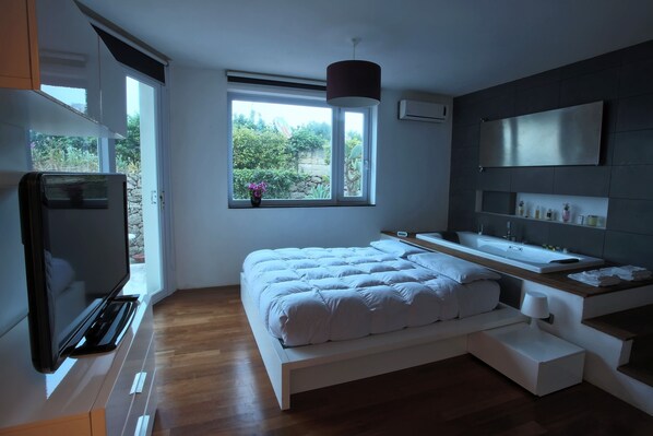 Un dormitorio moderno con una cama grande, televisor, ventana con vistas a zonas verdes, lavamanos integrado y espejo.