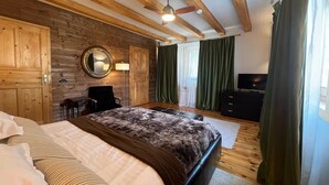 Un dormitorio con cama, una silla, un televisor, paredes de madera, ventilador de techo y cortinas.