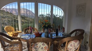 A dining area with wicker chairs, a table set for two, and a view of a mountainous landscape through large windows. - Rural villa with private outdoor pool, separate annexe and vast outdoor spaces (Gata De Gorgos)