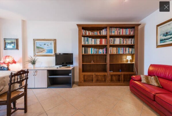 A living room with a red sofa, a wooden bookshelf, a television, and a dining area with a table and chairs.