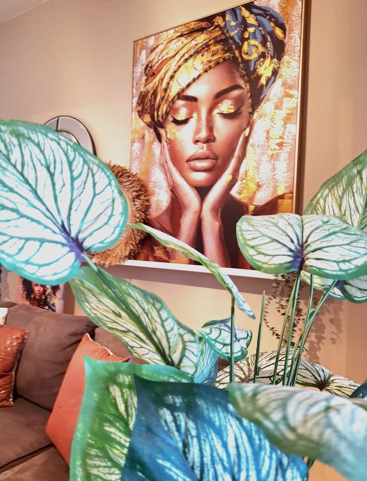 A room with a large painting on the wall, a sofa with cushions, and a potted plant with large leaves.