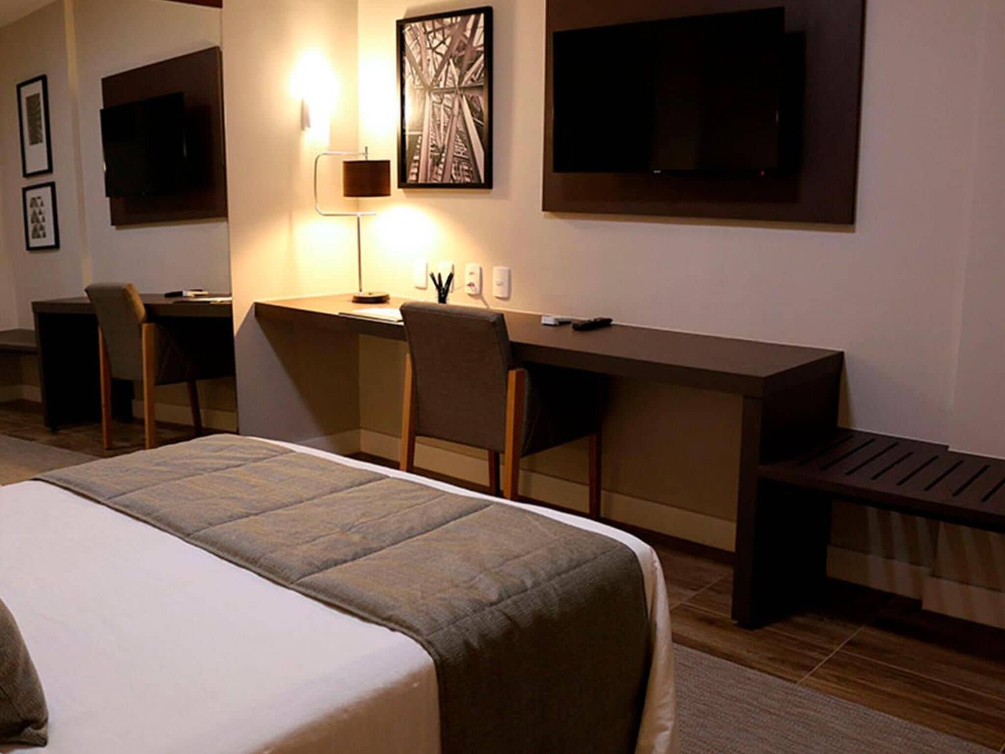 A hotel room with a bed, desk, chairs, a TV, and artwork on the wall.