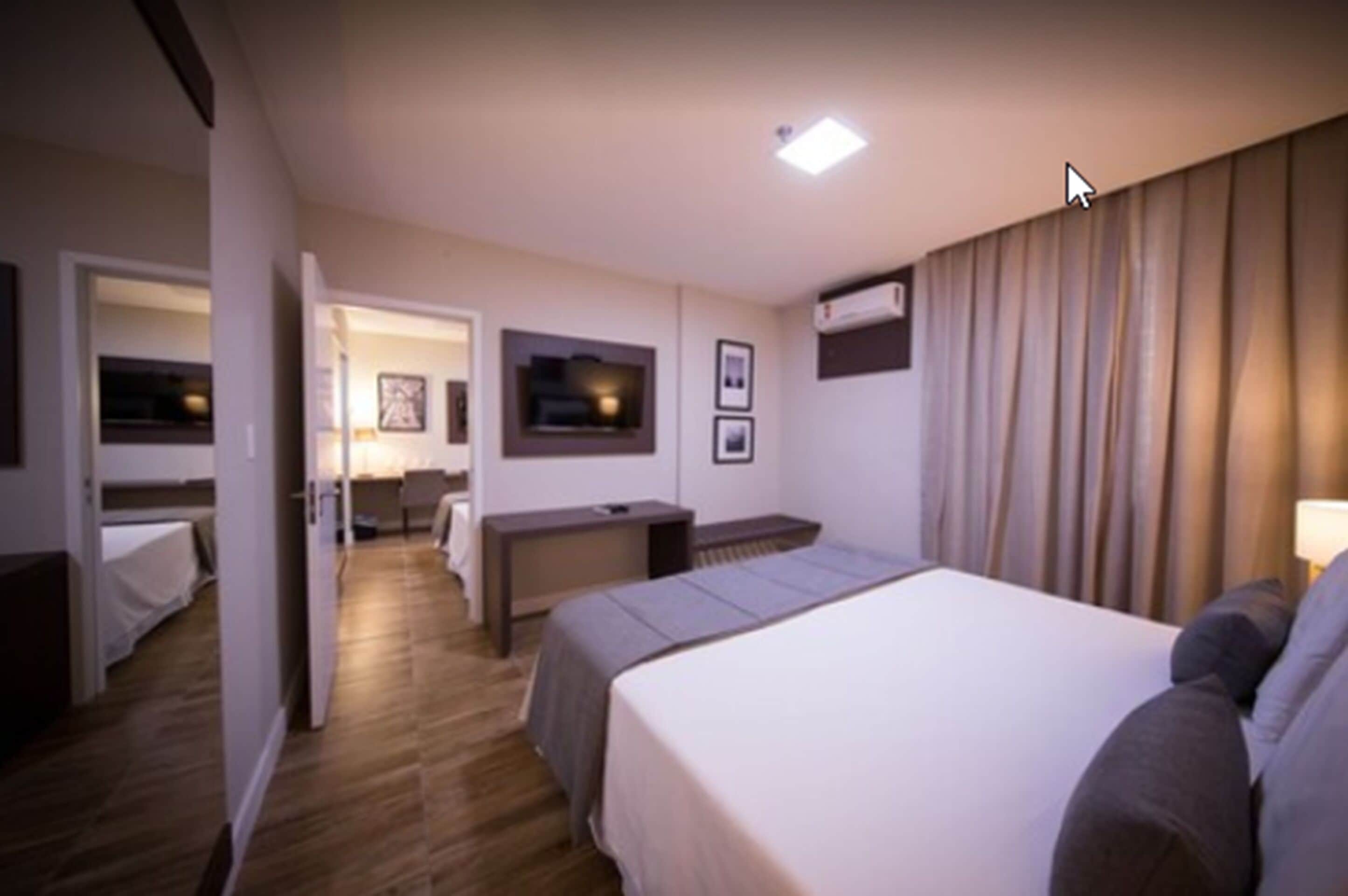A hotel room with a bed, bedside table, and a desk with a chair. There is a TV mounted on the wall and an air conditioning unit.