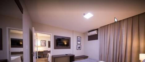 A hotel room with a bed, bedside table, and a desk with a chair. There is a TV mounted on the wall and an air conditioning unit.