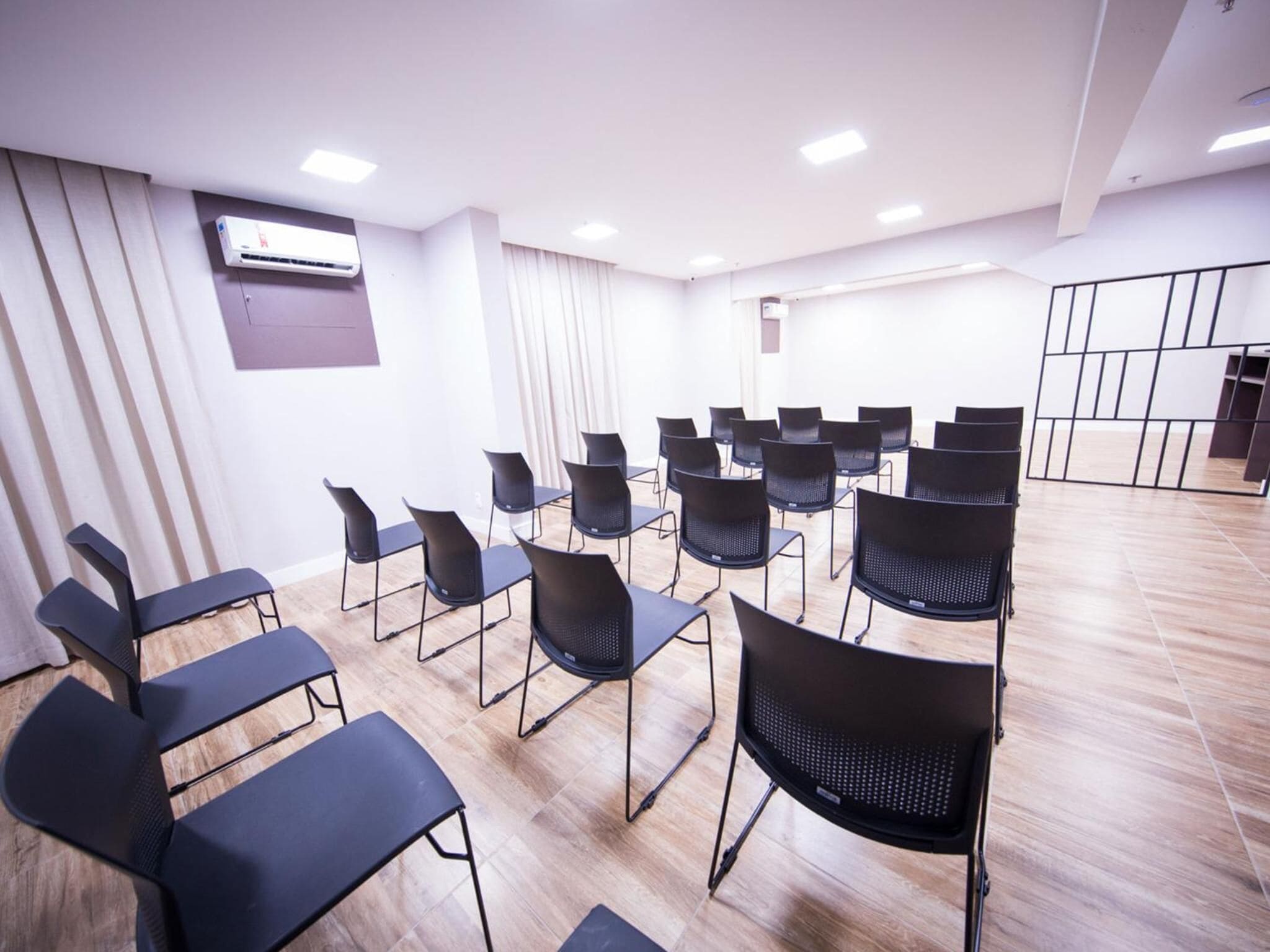 Un salón de conferencias con filas de sillas negras y mesas, suelo de madera, paredes blancas e iluminación empotrada en el techo.