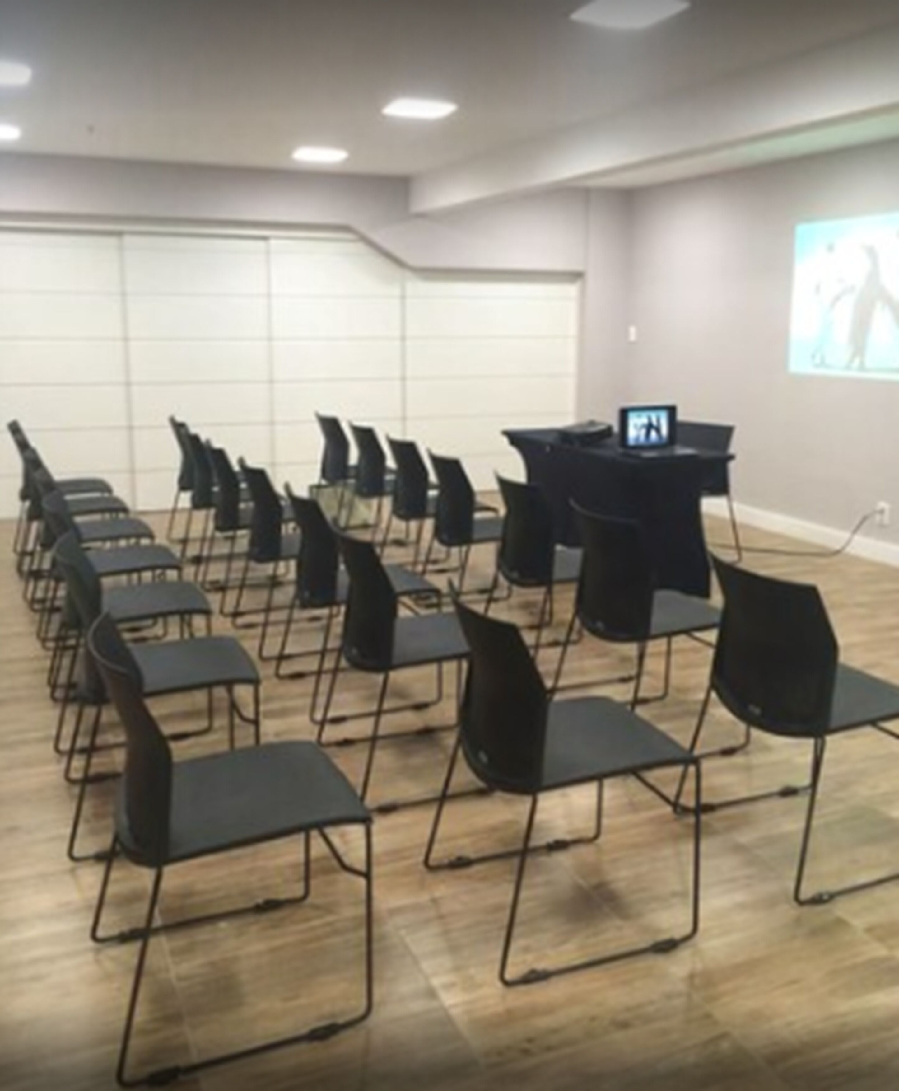 A conference room with rows of black chairs, a podium, and a screen displaying a presentation.