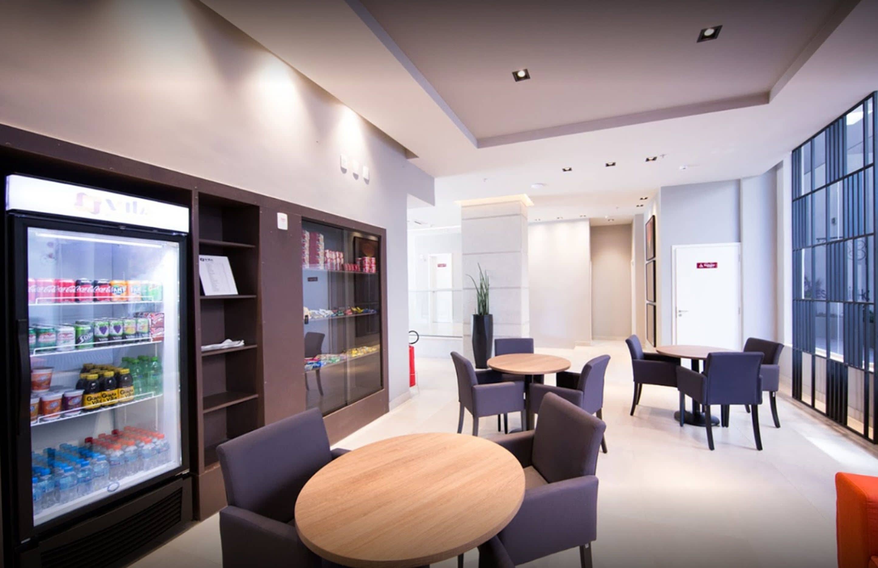 Un moderno hall de hotel con un expositor refrigerado de bebidas, zona de estar con mesas y sillas redondas, y amplias ventanas que ofrecen vistas.