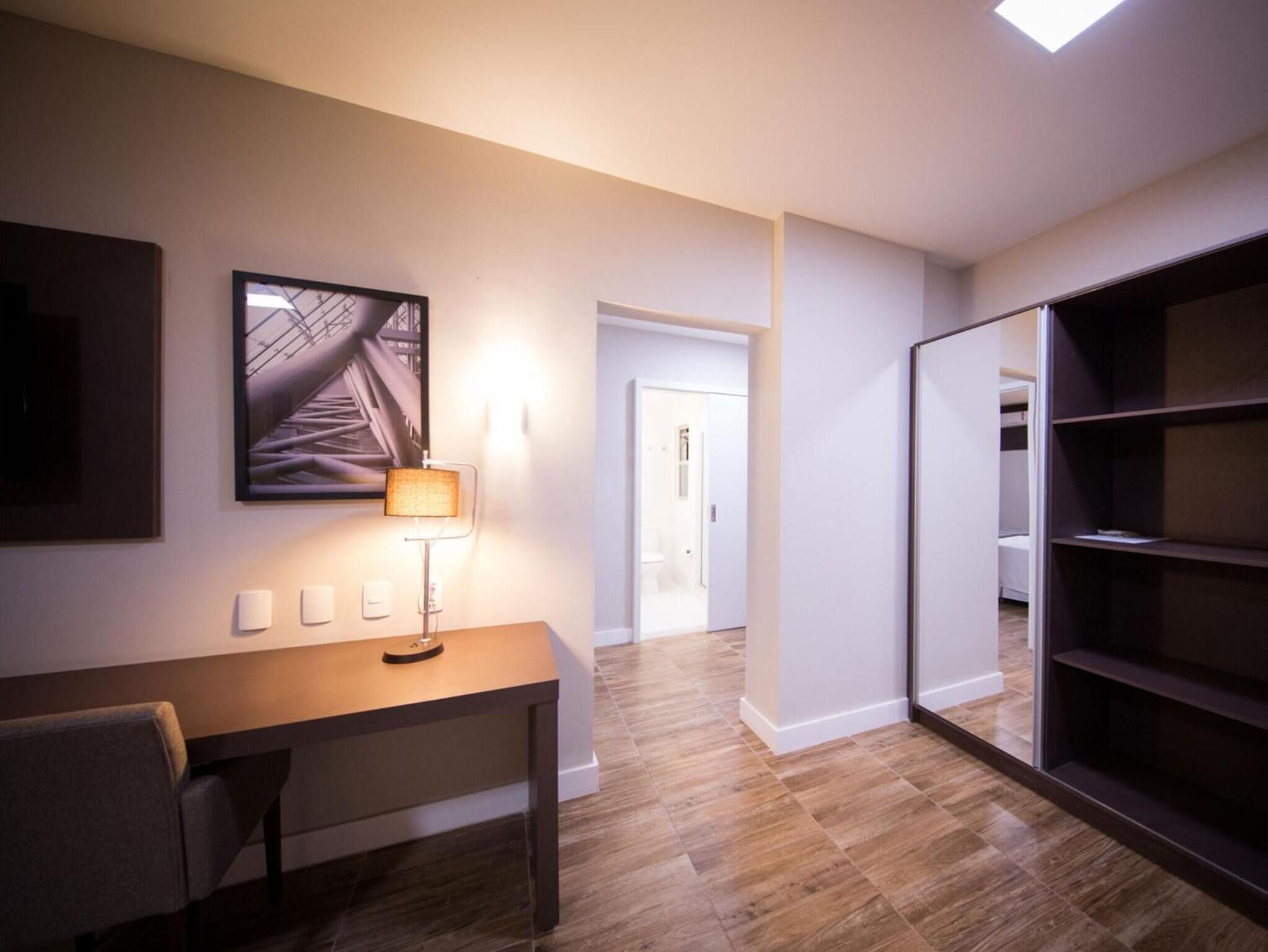 Una habitación de hotel moderna con piso de madera, un escritorio con lámpara, una cama y un armario.
