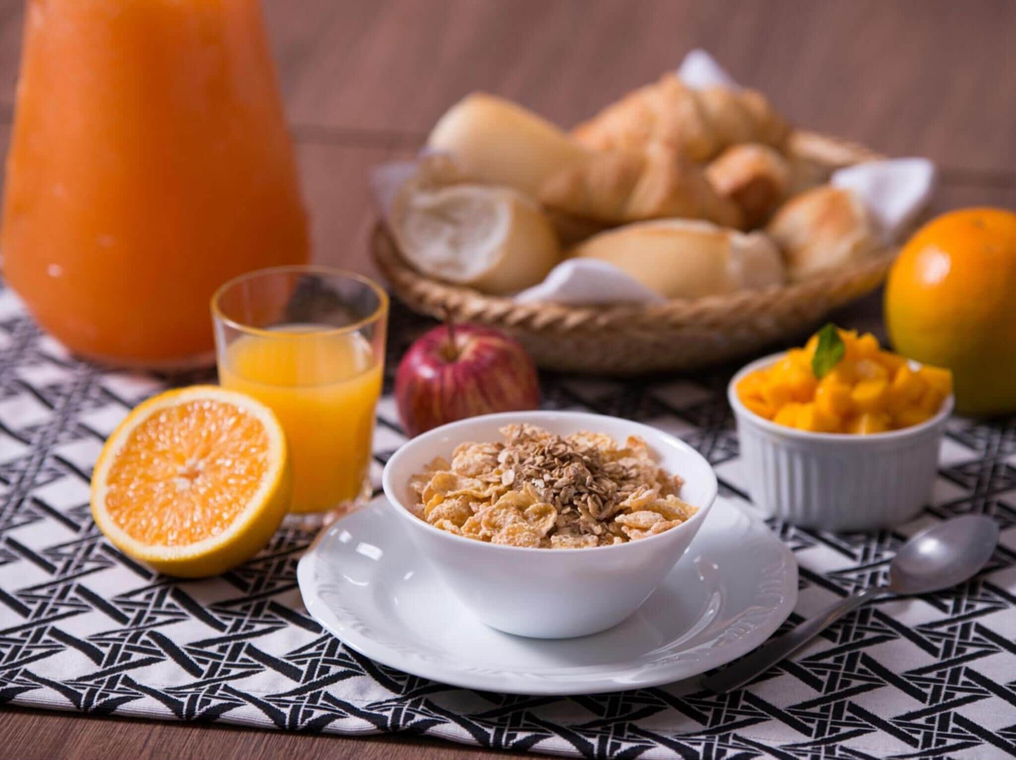 Un desayuno con un tazón de cereal, un vaso de jugo de naranja, una cesta de pan, una manzana y un tazón de trozos de mango sobre un mantel estampado.