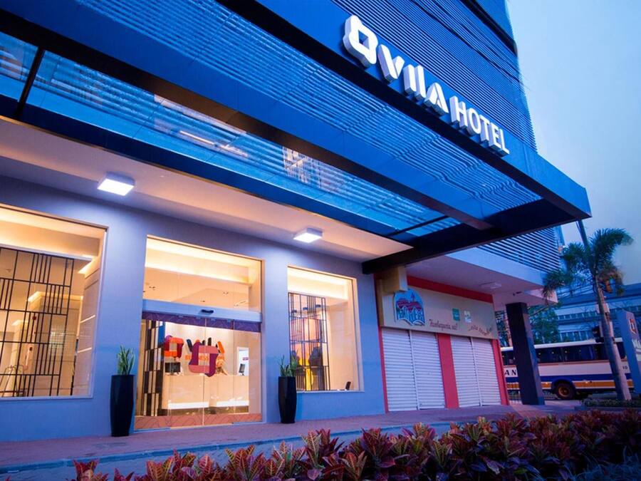 Exterior del Vila Hotel con un diseño moderno, destacando amplias ventanas de vidrio y un cartel llamativo.