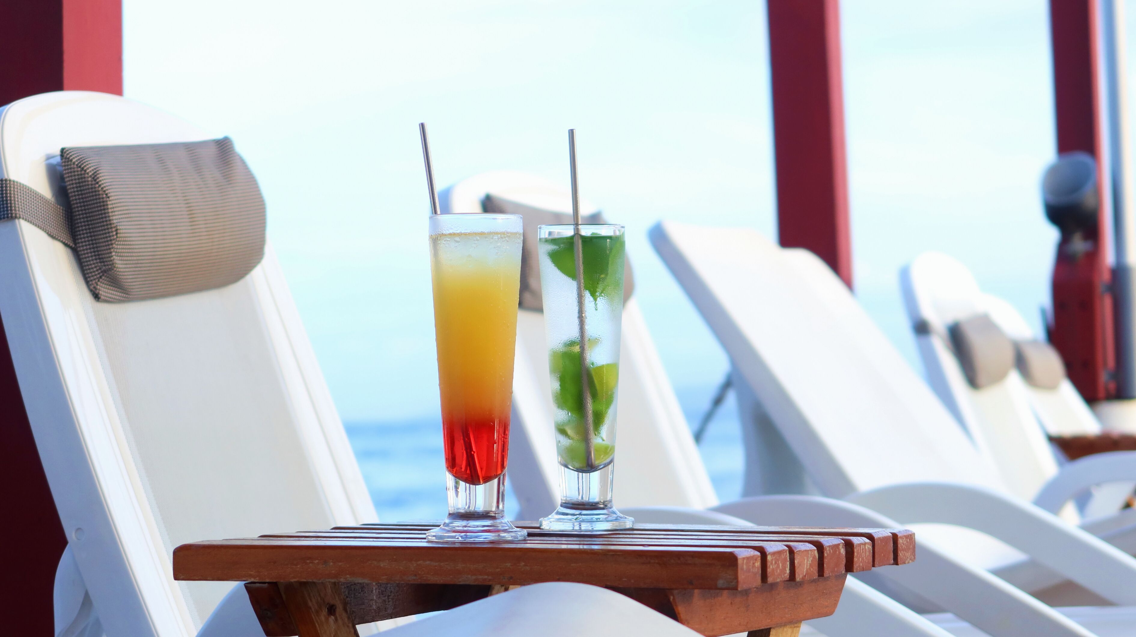 Two drinks on a wooden tray between white lounge chairs with a view of the ocean.