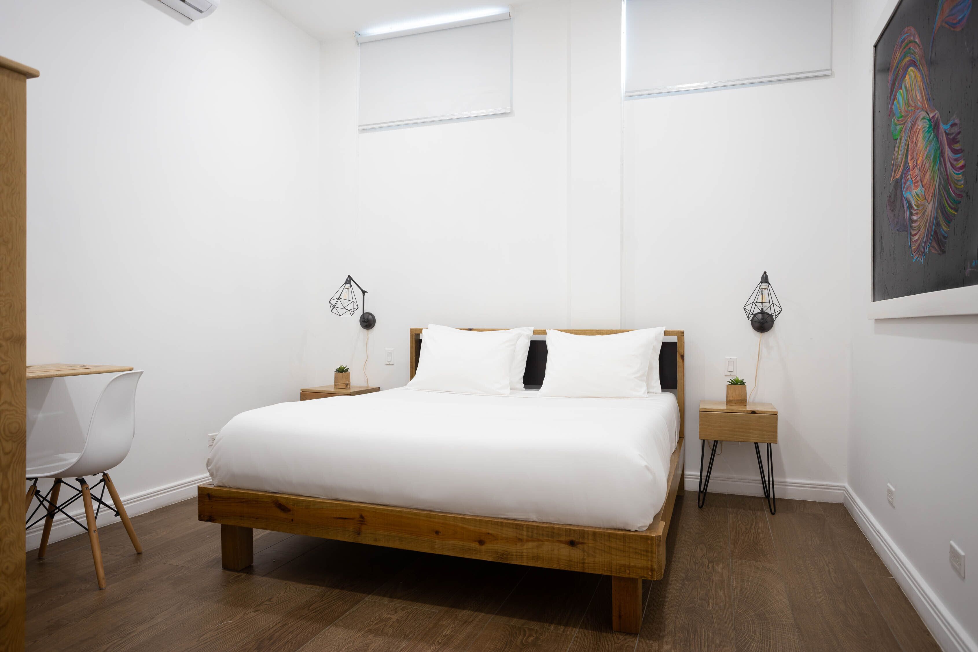 Un dormitorio con una cama de madera, ropa de cama blanca, una silla blanca y una mesita con una planta.