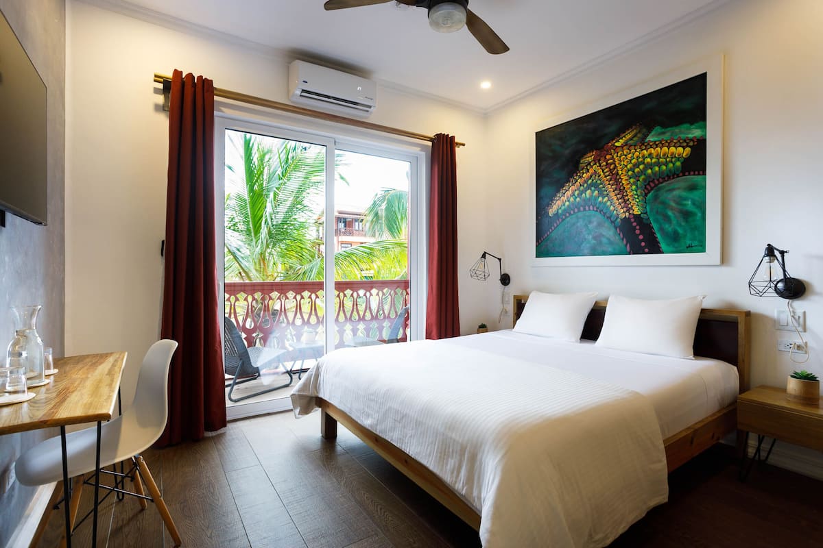 Ein Hotelzimmer mit Bett, Schreibtisch mit Stuhl, Fenster mit Vorhängen, Deckenventilator und einem Bild an der Wand.