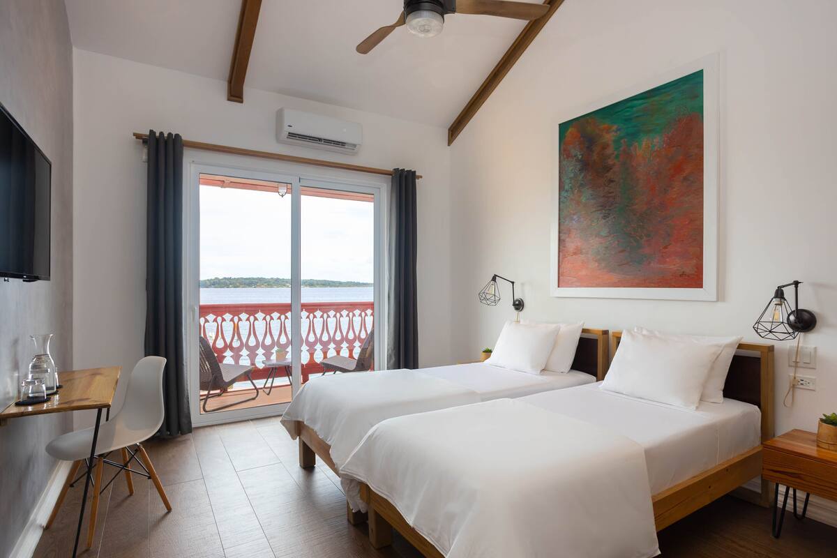Ein Hotelzimmer mit zwei Betten, einem Schreibtisch mit Stuhl, einem Balkon mit Aussicht und einem Bild an der Wand.