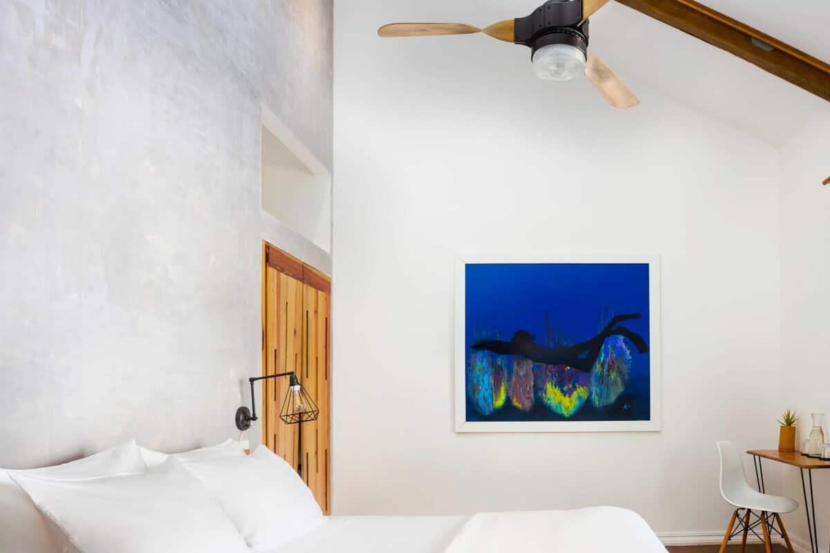 Ein Schlafzimmer mit einem Bett, einem Deckenventilator, einer Wandleuchte, einem Gemälde, einem kleinen Tisch mit Stuhl und einer Holztür.