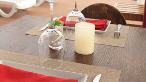 Mesa de jantar posta com taça de vinho, vela e pratos, com vista para uma praia com cadeiras de praia e uma pérgola de madeira.