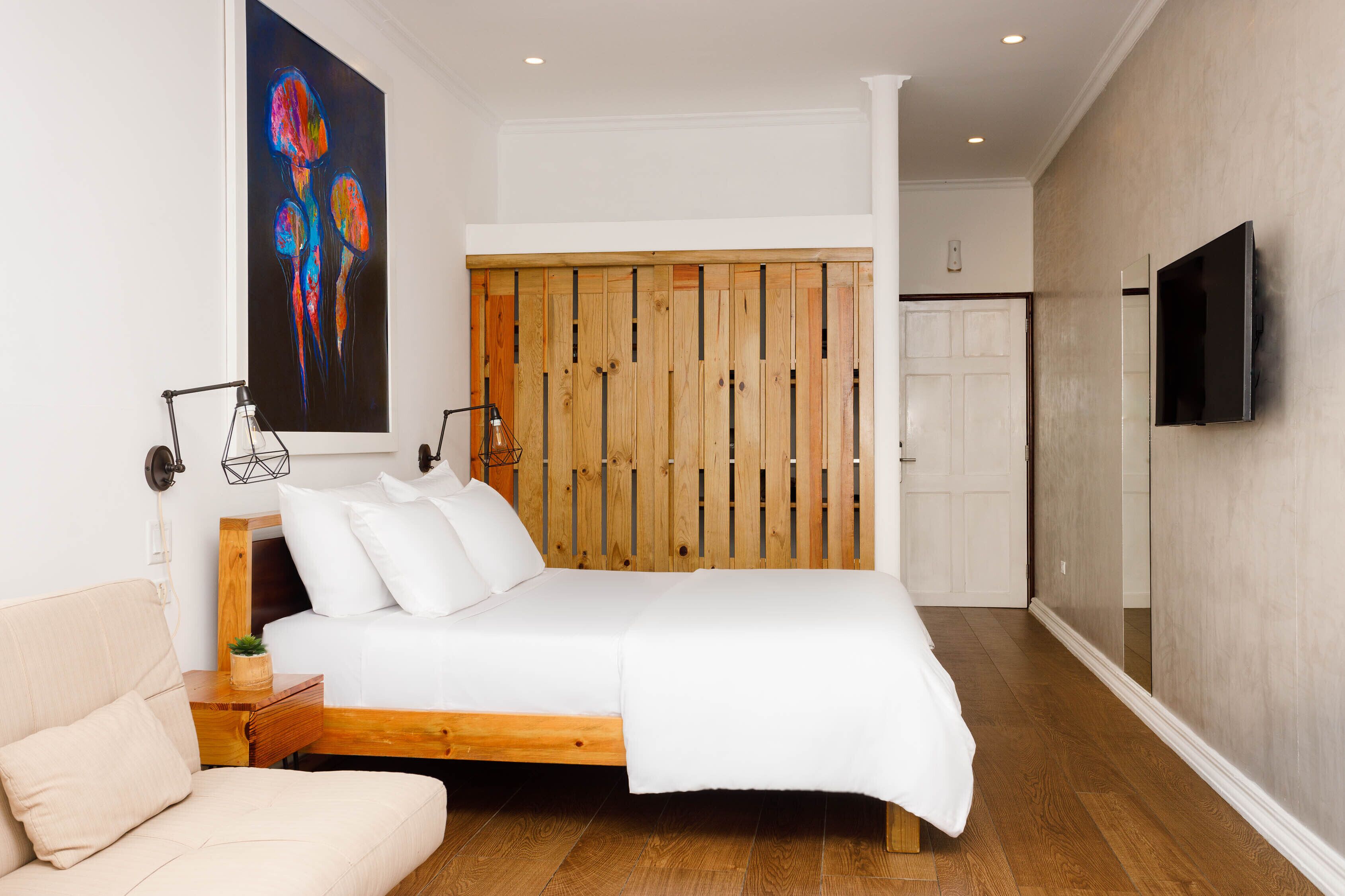 A bedroom with a wooden headboard, a bed with white bedding, a bedside table with a lamp, and a large abstract painting on the wall.