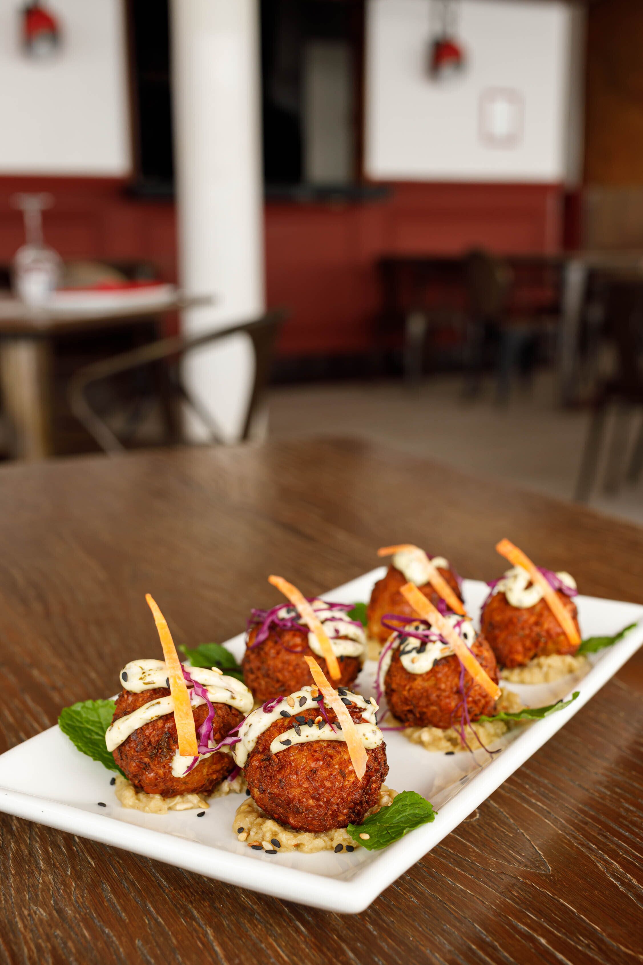 A plate of falafel balls on a white rectangular plate, garnished with vegetables and a white sauce, placed on a wooden table in a restaurant setting.