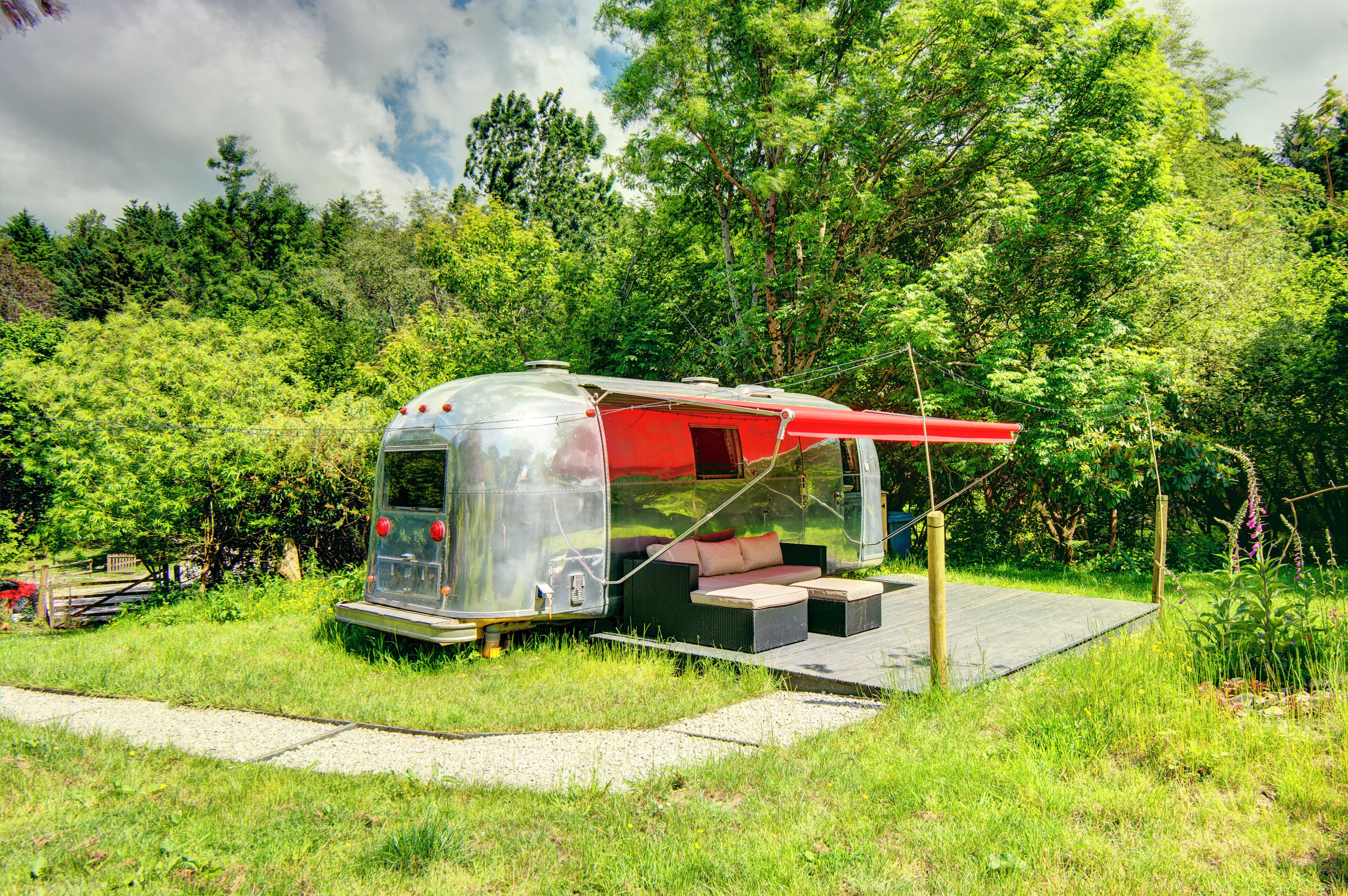 Una autocaravana transparent amb un toldo vermell, un sofà i una taula petita, situada en una zona verda amb arbres al fons.
