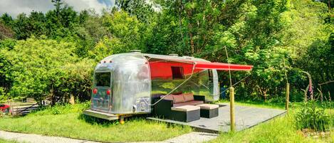 Una autocaravana transparent amb un toldo vermell, un sofà i una taula petita, situada en una zona verda amb arbres al fons.