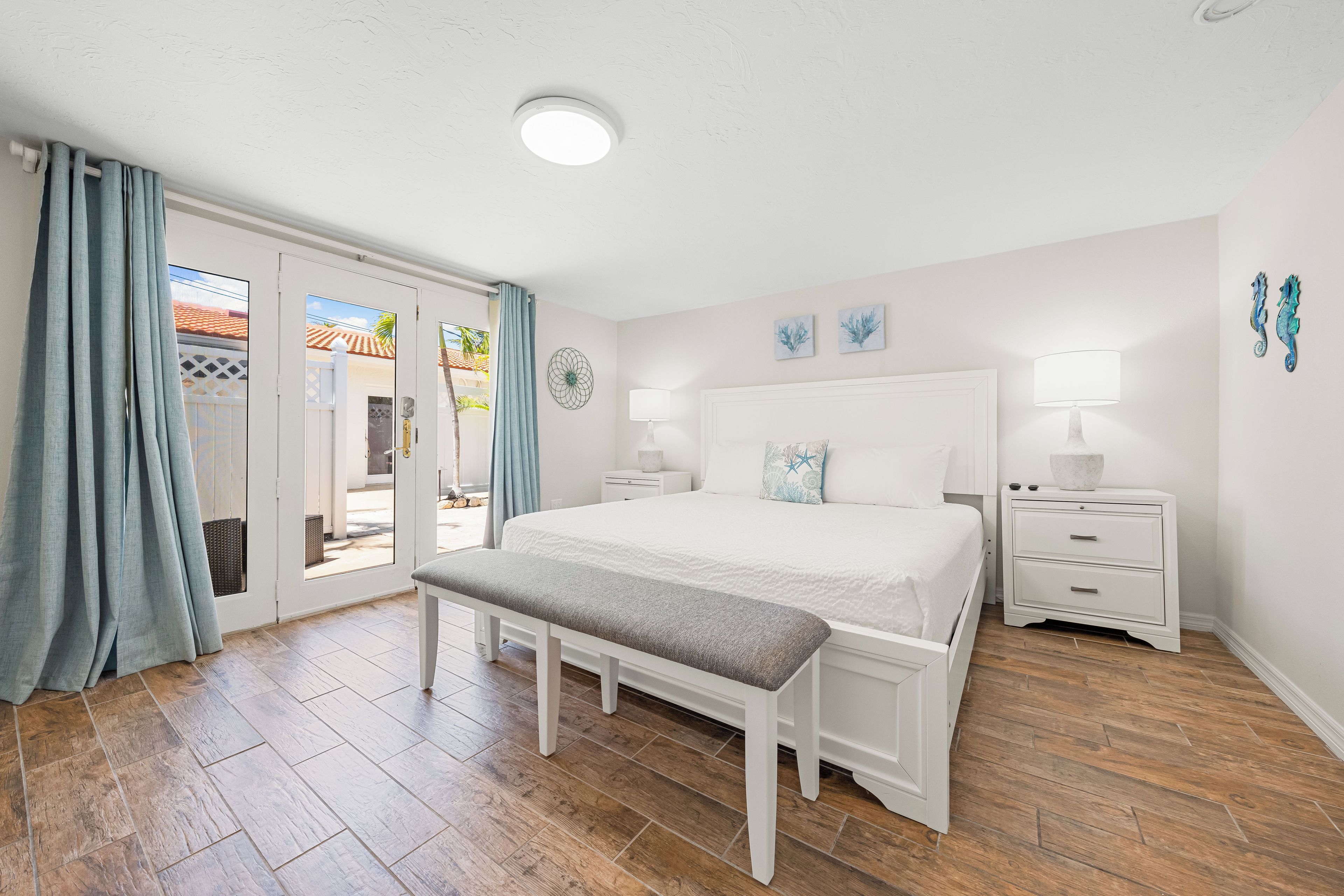 Quarto com uma cama grande, um banquinho, uma mesa de cabeceira e vista para o exterior.