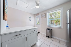 A kitchen with white cabinets, a stainless steel refrigerator, a trash can, and a window with blinds. - Bianco Sands by Beachside Management (Siesta Key)