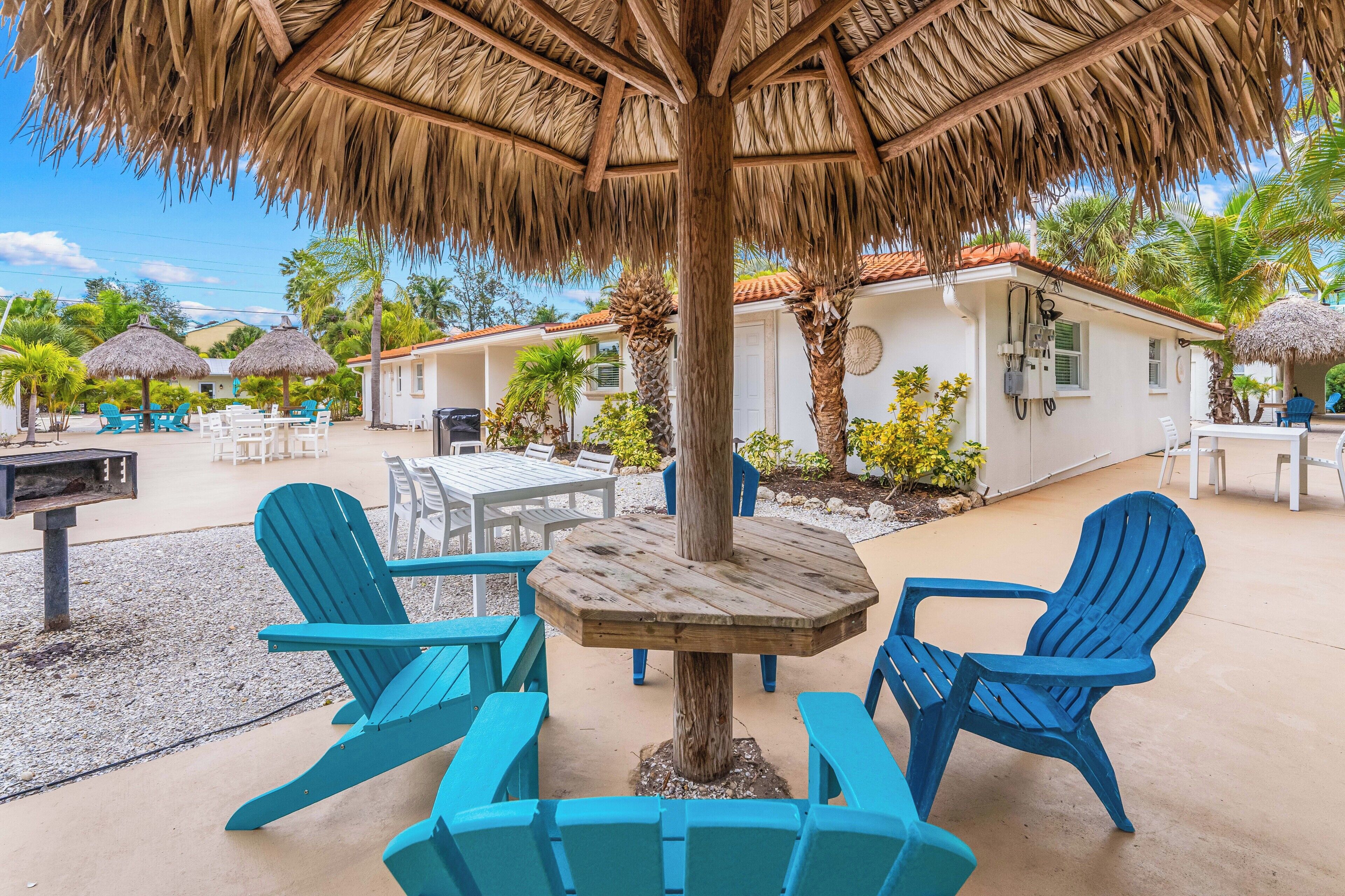 An outdoor seating area with a wooden table and blue chairs, surrounded by palm trees and a thatched roof.