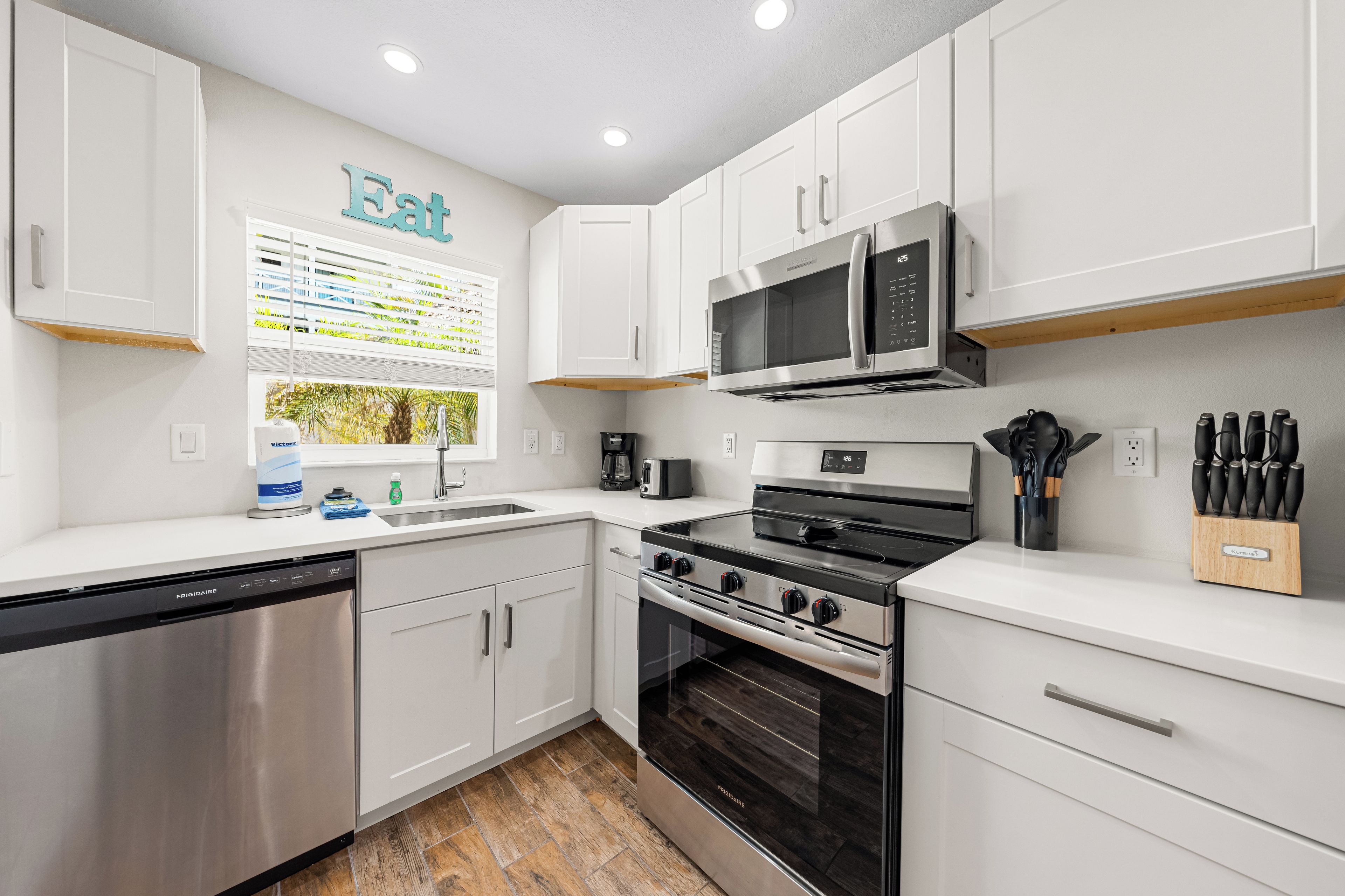 A modern kitchen with white cabinets, stainless steel appliances, and a window with a view of greenery.