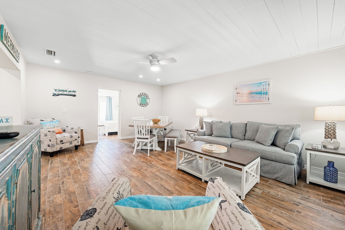 A spacious living room with a wooden floor, a grey sofa, a white dining table with chairs, and a ceiling fan.