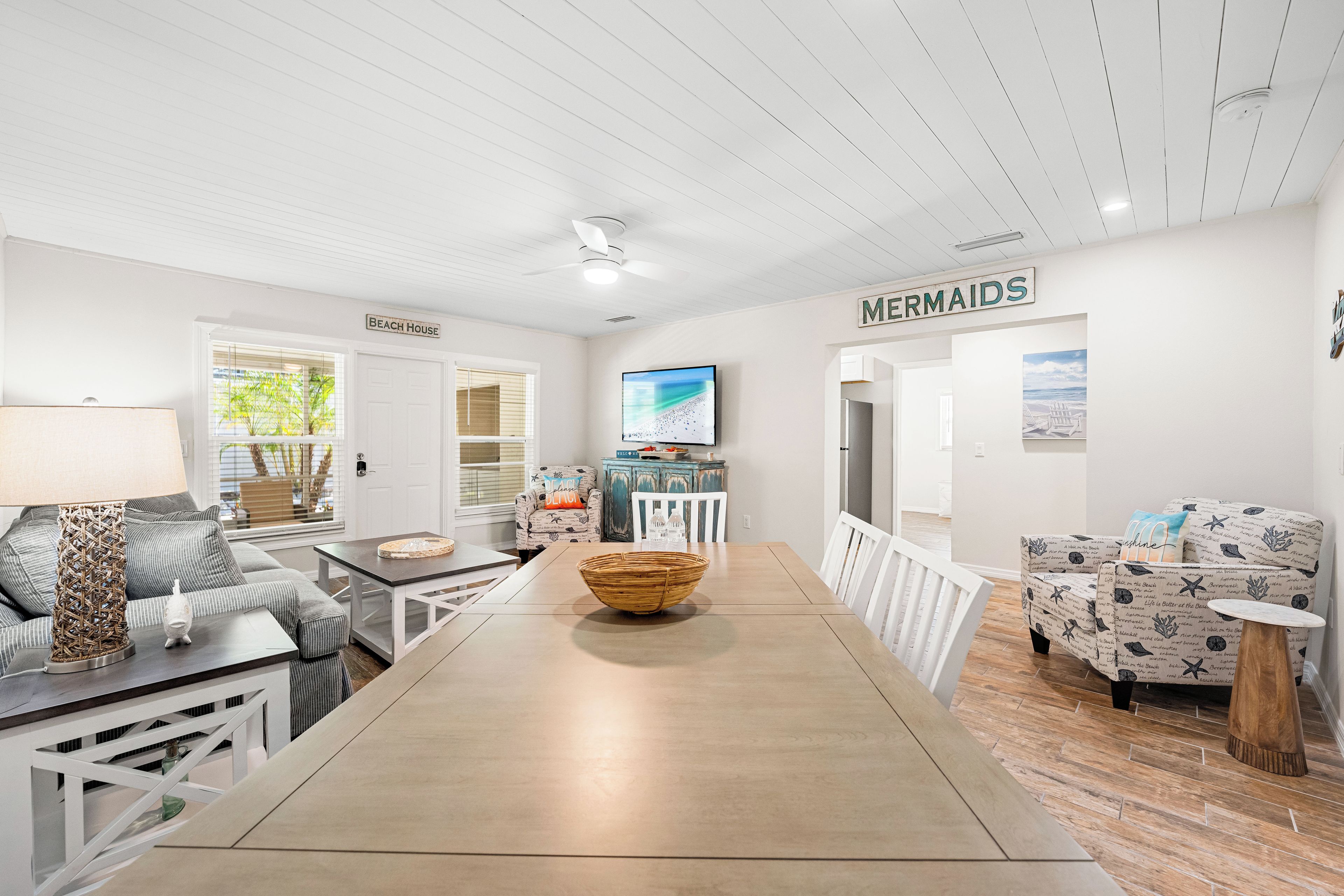 Uma sala de estar moderna com uma mesa de jantar, um sofá e uma TV. Há um letreiro na parede que diz "MERMAIDS".