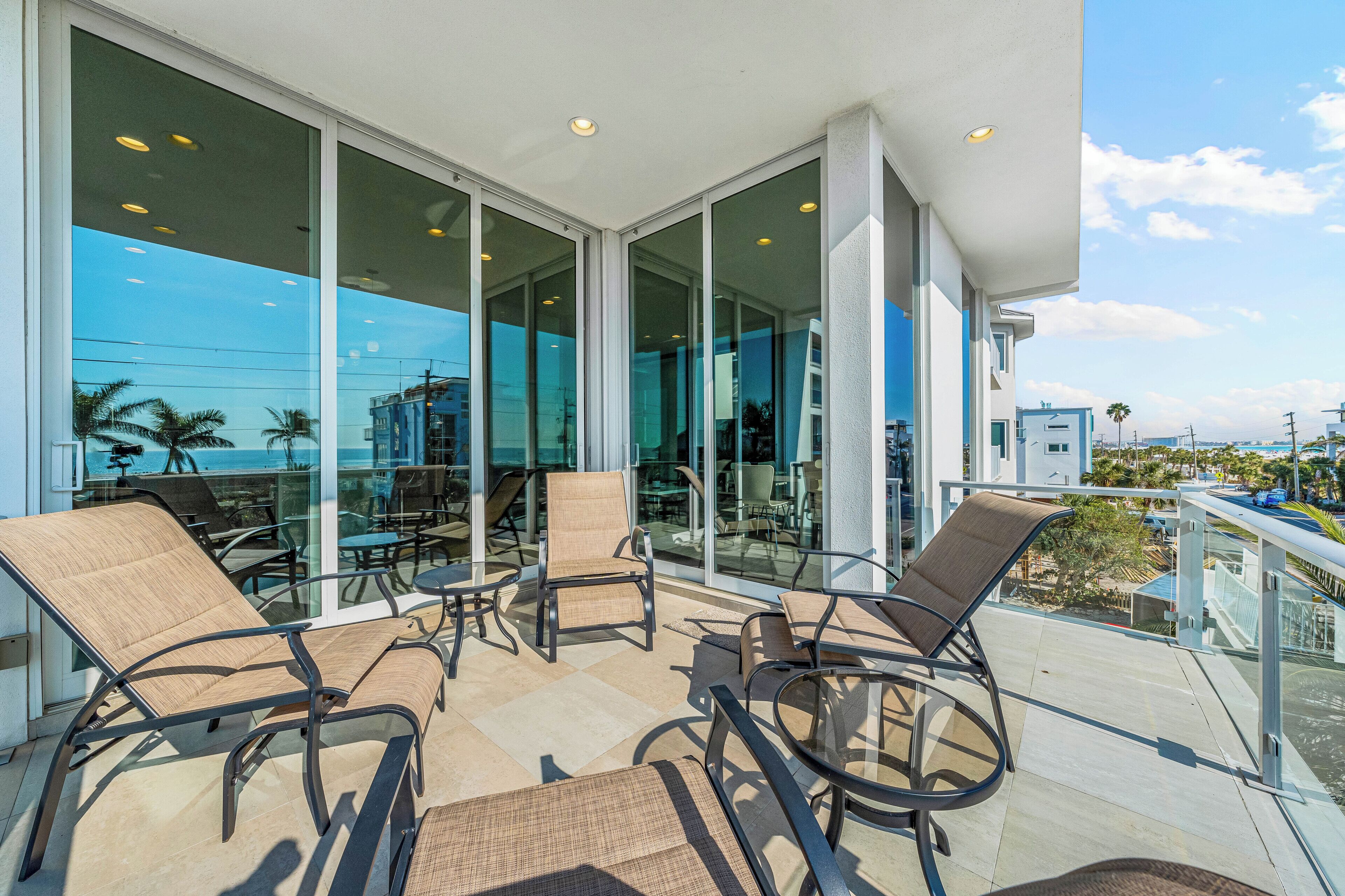 A balcony with outdoor seating, large glass windows, and a view of the ocean and buildings.