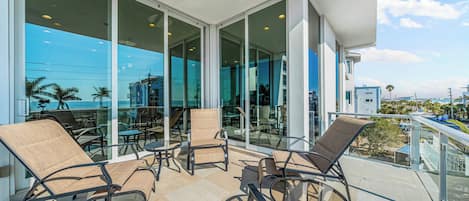 A balcony with outdoor seating, large glass windows, and a view of the ocean and buildings.