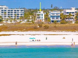 A beach with clear turquoise water, white sand, and several buildings in the background.