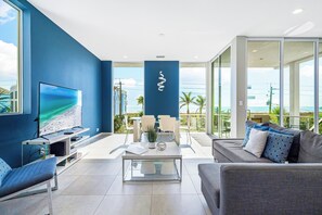 A modern living room with a large TV, a sofa, a coffee table, and a balcony with a view of the ocean.
