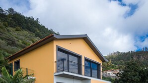 A modern two-story house with a balcony, a patio with chairs, and a grassy yard. - Gorgeous, Modern Home with Breathtaking Views (Arco da Calheta)