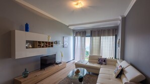 A modern living room with a flat-screen TV, a glass coffee table, a beige sofa, and a wooden entertainment unit. - Gorgeous, Modern Home with Breathtaking Views (Arco da Calheta)