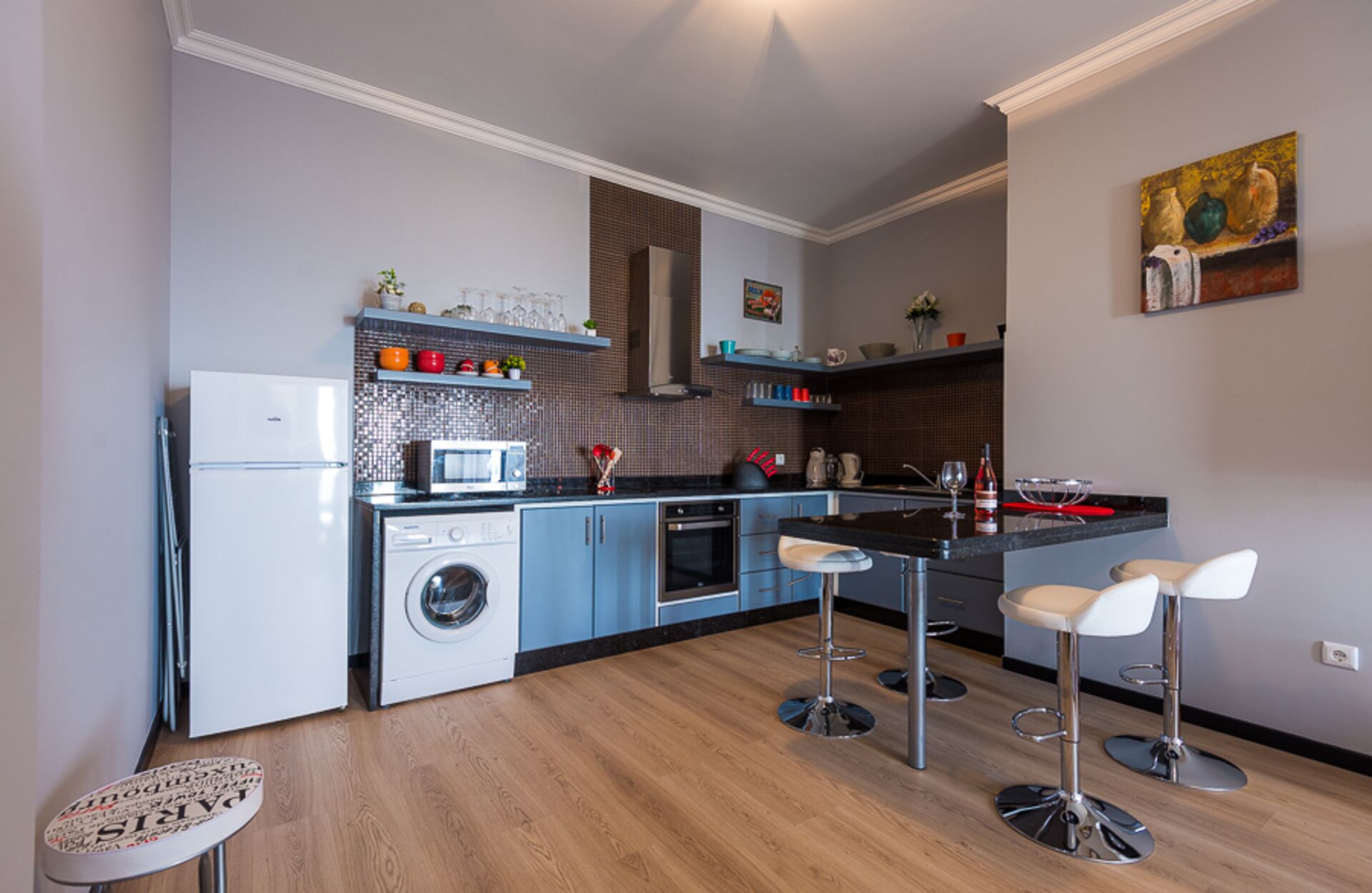 Een moderne keuken met een wasmachine, magnetron en oven. Er zijn barkrukken en een klein tafeltje met een decoratief object.