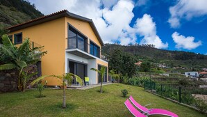 A modern two-story house with a yellow facade, a balcony, and a grassy yard with pink chairs. - Gorgeous, Modern Home with Breathtaking Views (Arco da Calheta)